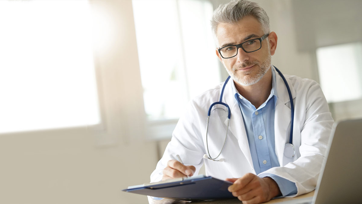 Image of an older White man doctor writing on a navy blue clipboard and sitting in a bright white and beige office